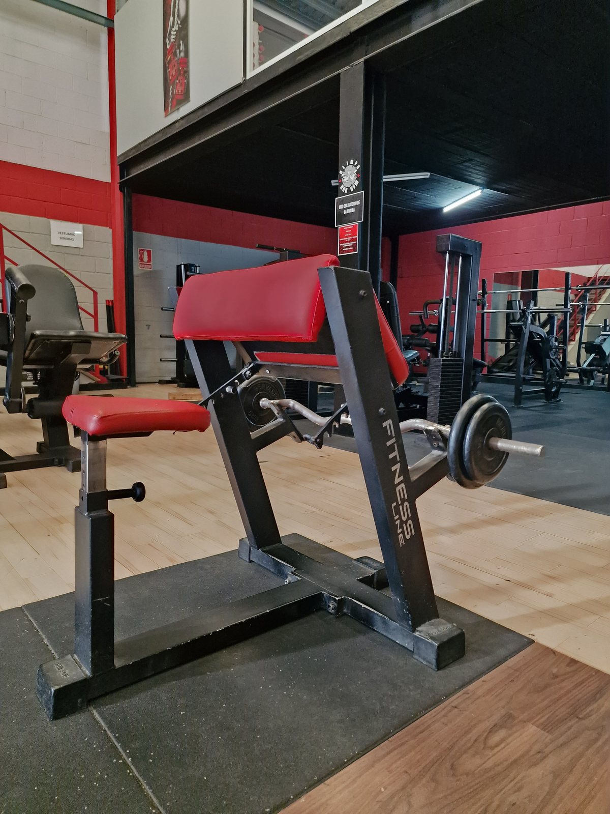 Un banco de curl predicador con tapicería roja y un barbell en un gimnasio con paredes rojas y suelo de madera.