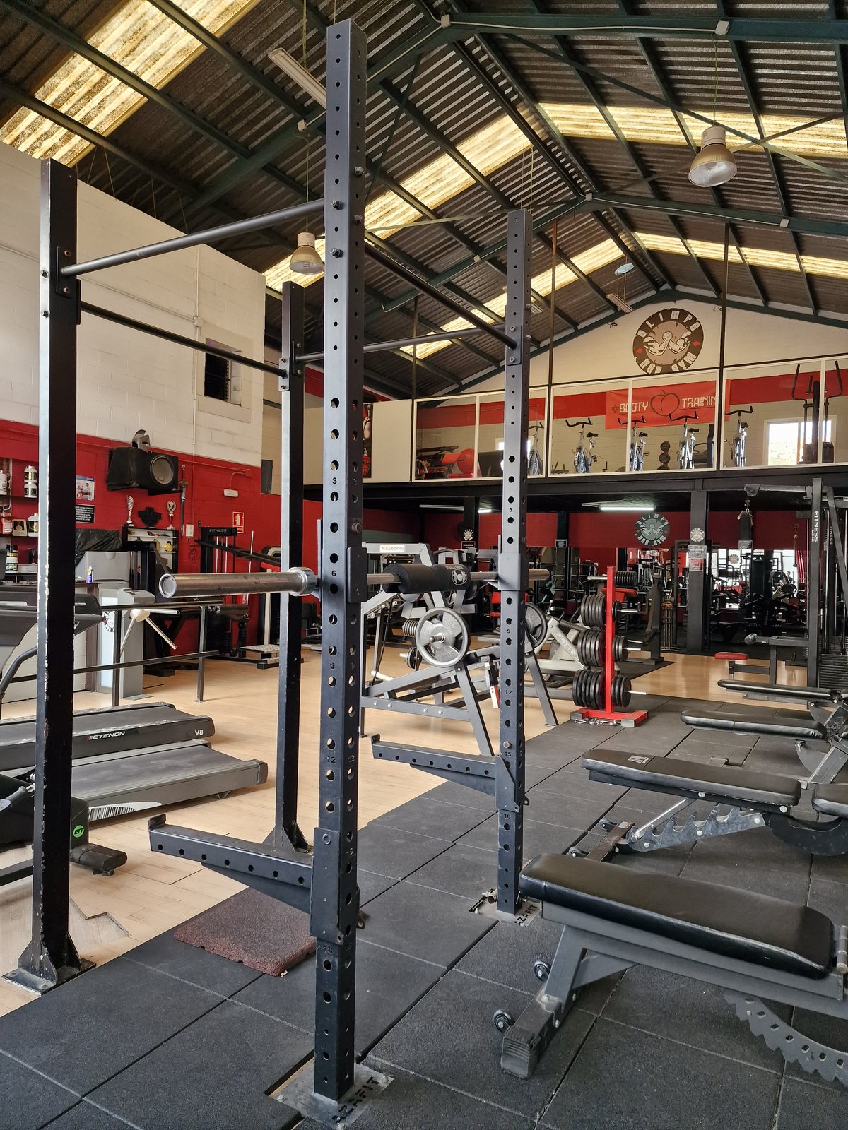 Interior del gimnasio Olimpo NGR con una jaula de sentadillas y bancos en primer plano, y cintas de correr y otras máquinas en el fondo.