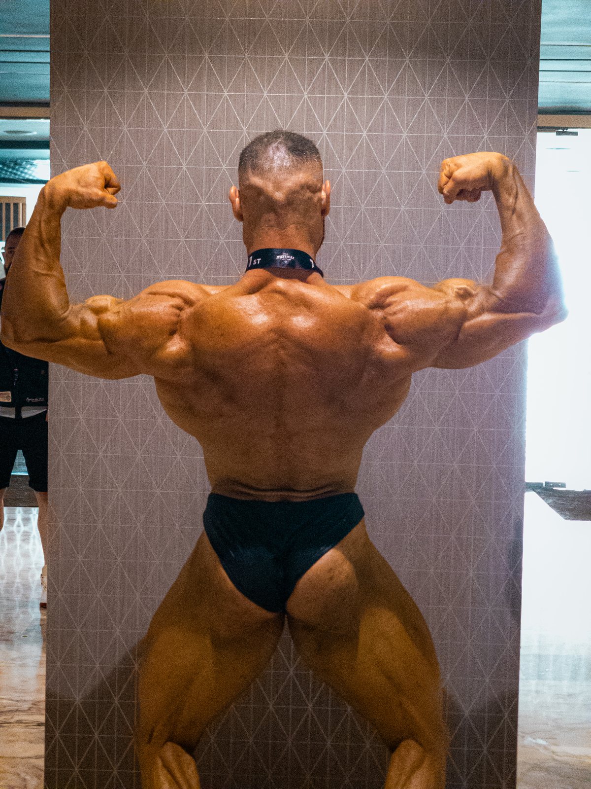 Un culturista musculado, con la espalda bronceada, posando de espaldas con doble bíceps, lleva un slip negro y un collar.