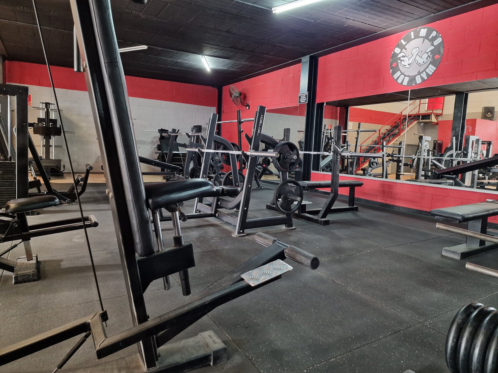 Interior del gimnasio Olimpo NGR Gym, mostrando una variedad de máquinas de musculación y pesas libres, con paredes rojas y negras.