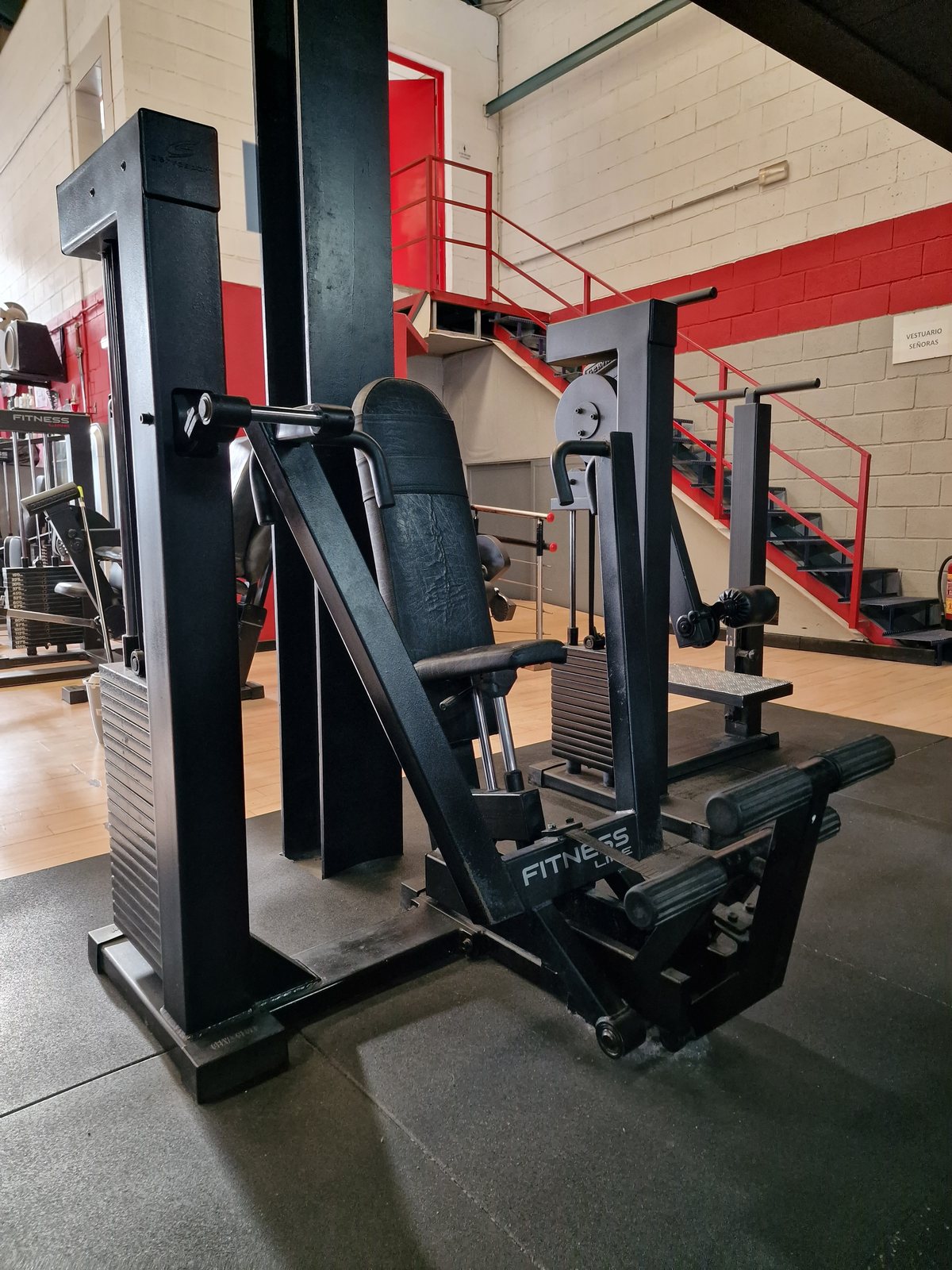 Una máquina de prensa de piernas y otra máquina de gimnasio en el interior de un gimnasio, con escaleras rojas al fondo.