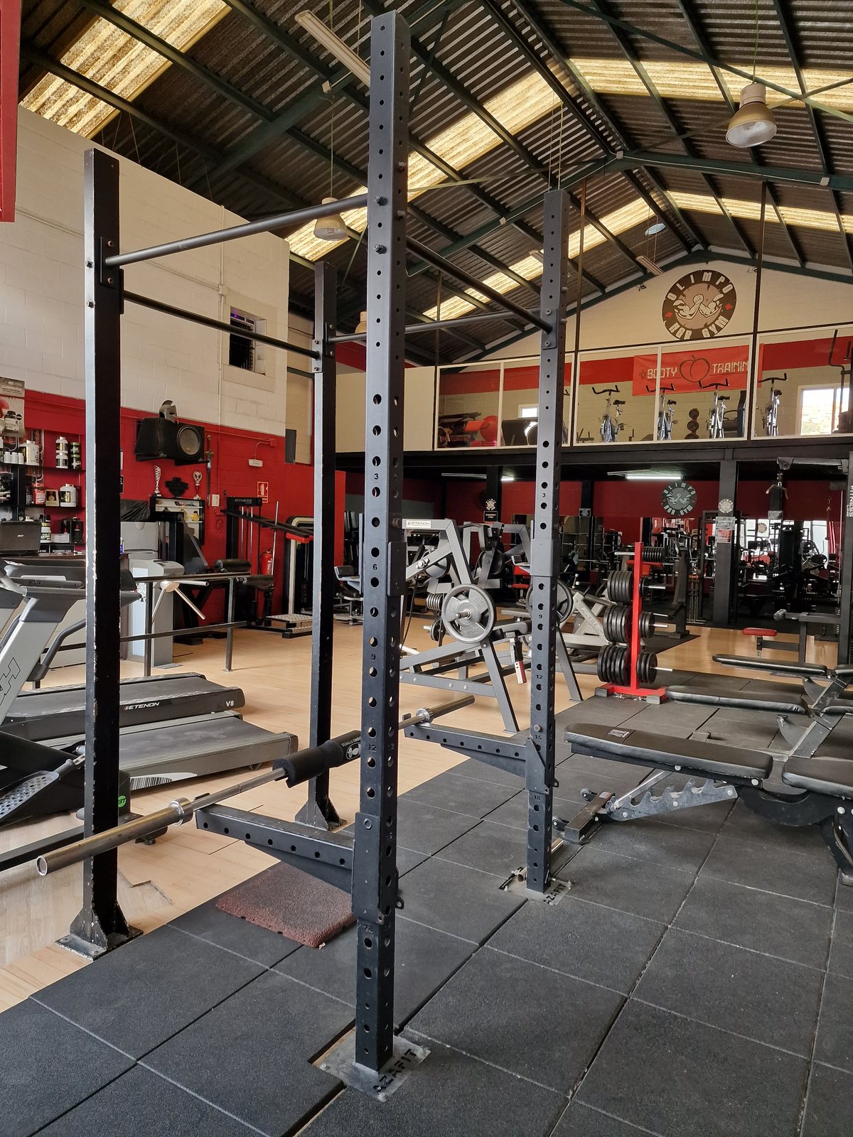Interior de un gimnasio con una jaula de sentadillas en primer plano y diversas máquinas de entrenamiento y peso libre al fondo.